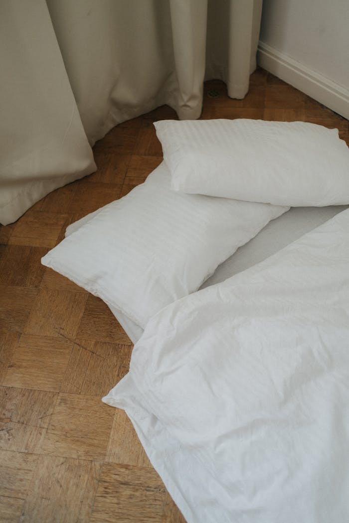 A simple minimalist setup with white bedding and pillows on a wooden floor, emphasizing simplicity and calm.